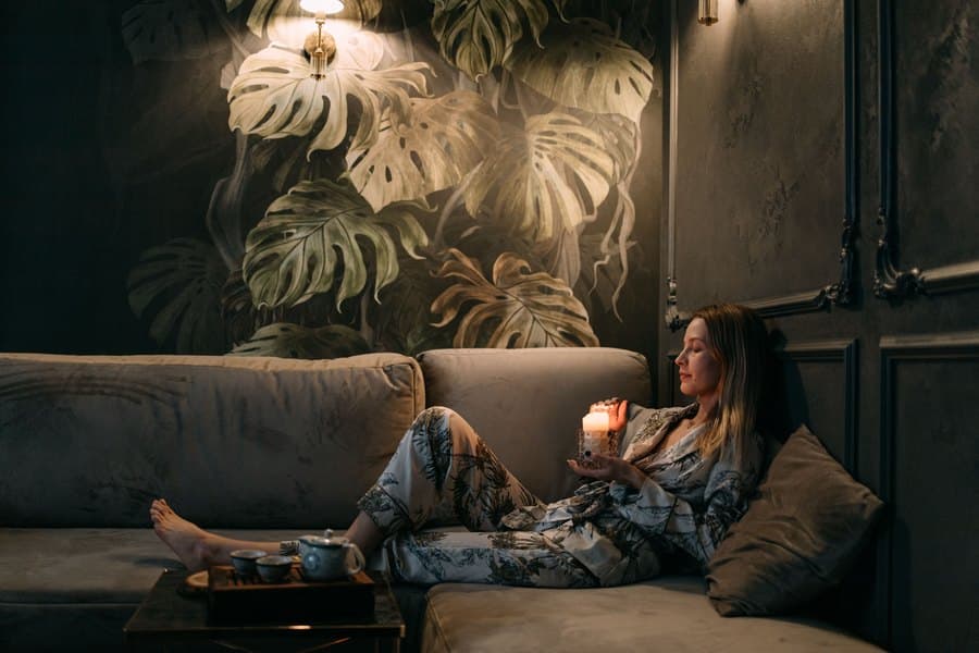 Woman resting on a sofa by candlelight against a dark botanical wall